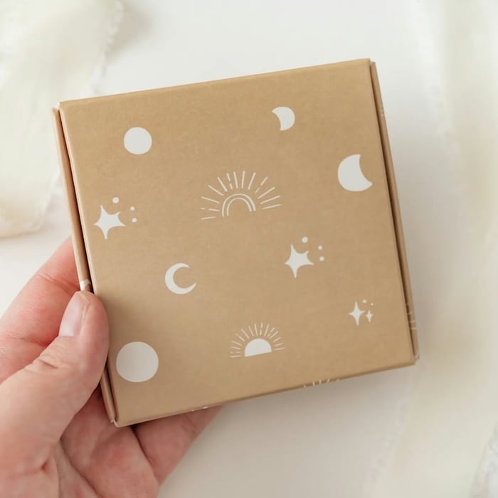 A small kraft box printed with white moons, suns and stars, held in one hand